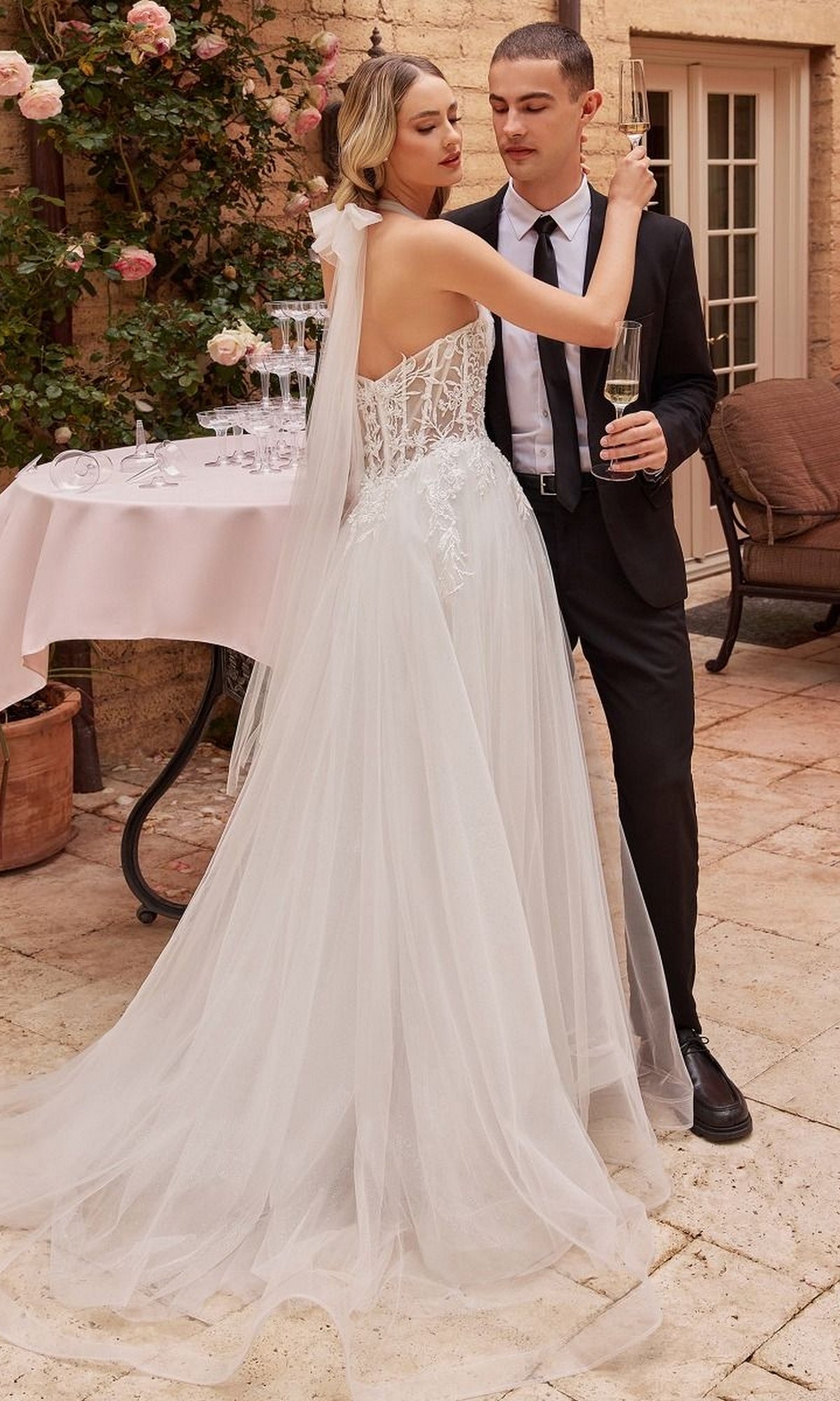 A bride in an Off White Long Halter Wedding Dress CD827W stands beside a groom in a black suit. They hold champagne glasses outdoors, with roses and a table visible in the background.
